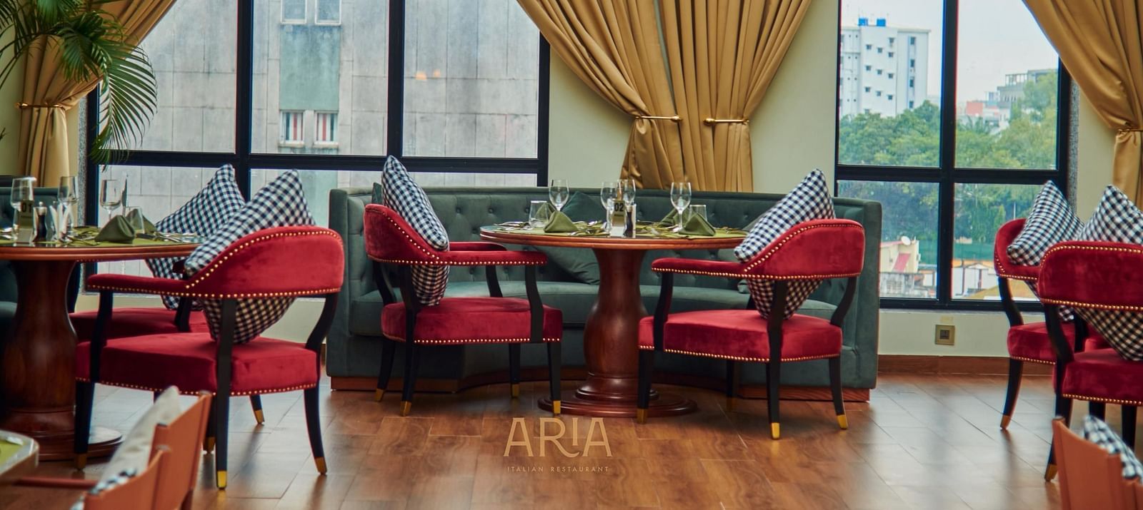 Elegant Aria restaurant interior with cushioned chairs, wooden tables, and large windows framed by curtains, allowing natural light to fill the space. City buildings and greenery are visible outside