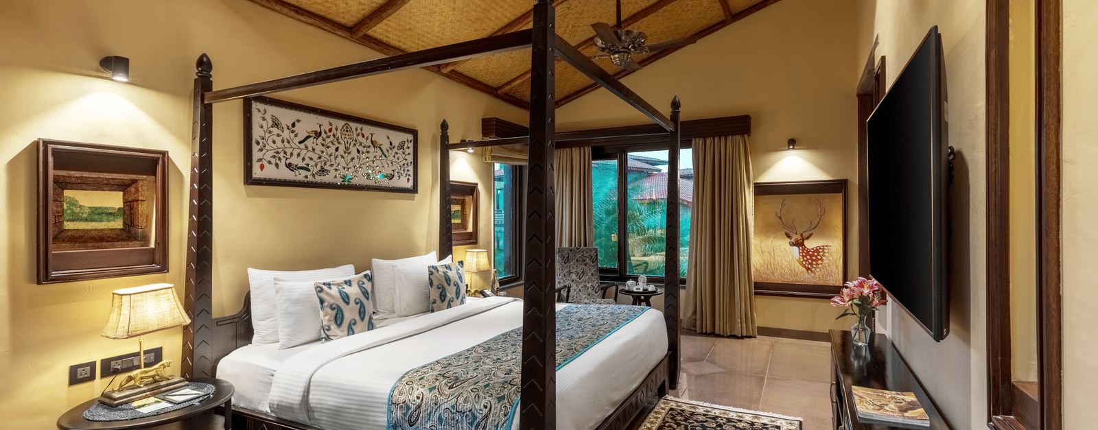 A luxurious four-poster bed room at Aamaghati Wildlife Resort with warm earthy tones, classic furniture, patterned rug and large windows opening to natural views.