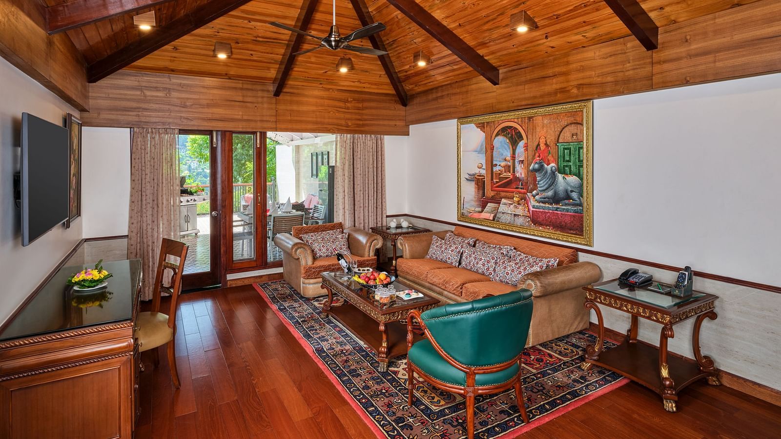A beautiful interior seating area of Imperial Villa with a table, sofas, chandelier, TV,  and a window overlooking greenery.