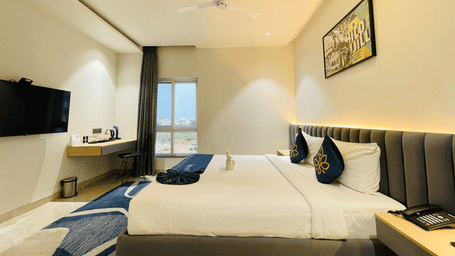  Hotel room at Hotel Kyzen HiTech City featuring a king bed with blue accent bedding, wall-mounted TV, framed artwork, ceiling fan, and large windows with natural light.
