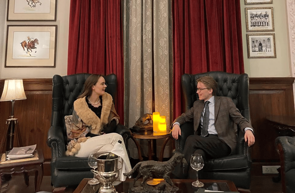 Two people sit across a coffee table having a conversation in a classic lounge with red curtains - Khas Bagh Jaipur.