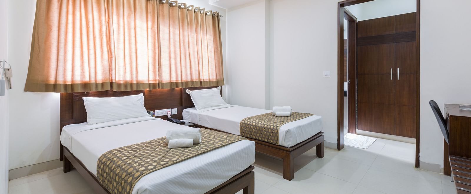 A room with twin beds featuring patterned bed runners and curtains, with an open doorway - Sanctum Suites BEL Road, Bangalore