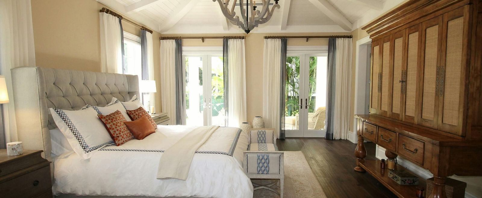 A spacious bedroom with a white bed, vaulted ceiling, chandelier, and a large wooden wardrobe.