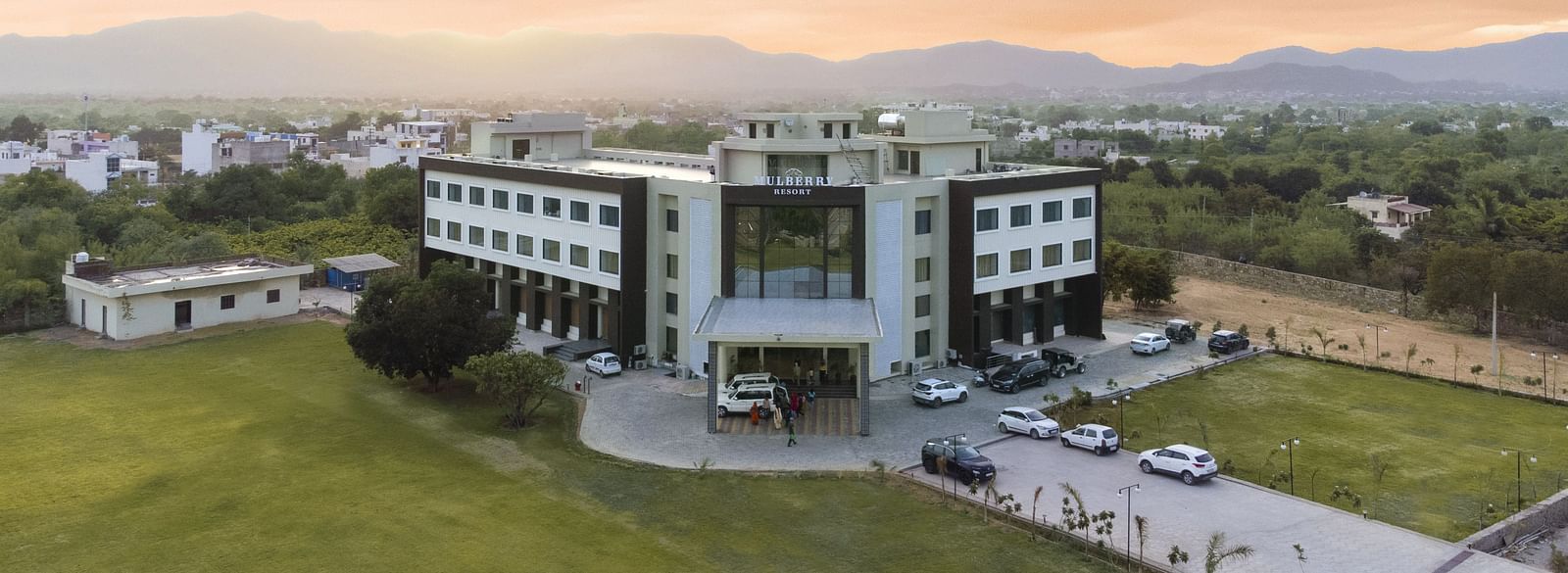 An aerial view of the Bamboo Saa Mulberry Resort, Pushkar at sunset or sunrise surrounded by green lawn and the backdrop of hills.