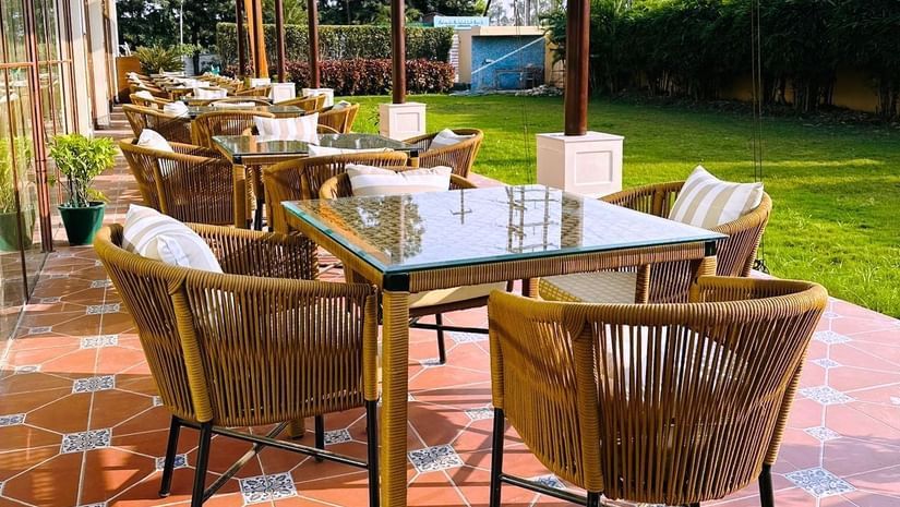  An outdoor patio with brown wicker chairs and glass-topped tables, shaded by a clear roof, overlooking a bright green lawn.