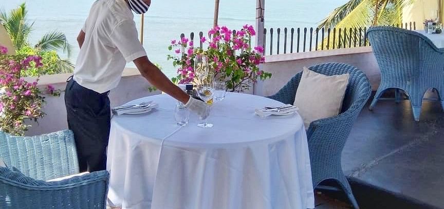 A staff member in a white uniform is setting or preparing an outdoor table at Fort Tiracol, Goa, with the ocean visible in the background through large windows or an open patio. 