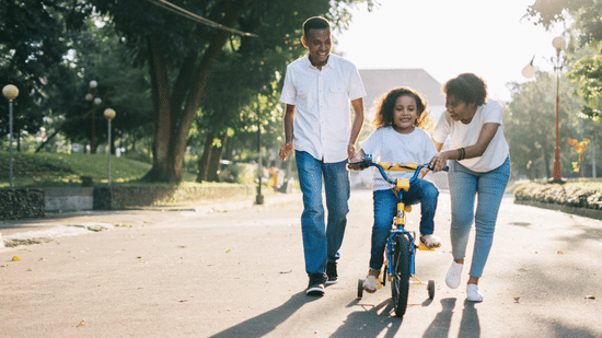 A mother and father smiling as they help their young daughter learn to ride a bicycle with training wheels on a sunny park path.