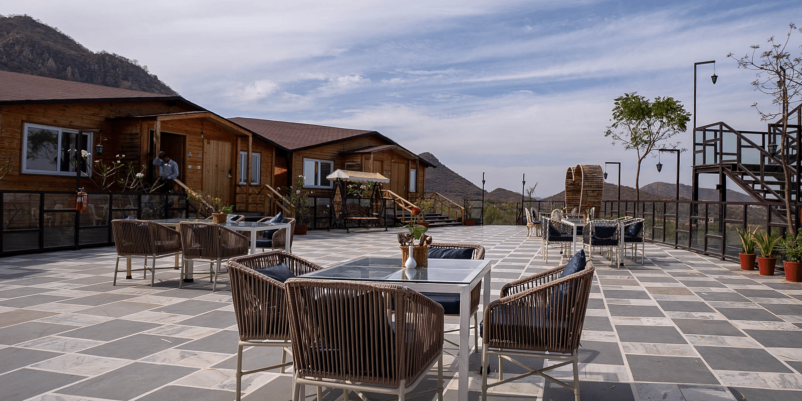 Outdoor patio with tables and chairs, wooden buildings in the background under a cloudy sky.- Bamboo Saa Resort & Spa, Udaipur