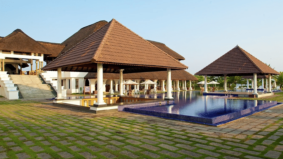 A paved courtyard beside an outdoor swimming pool, framed by thatched-roof resort pavilions | Le Pondy