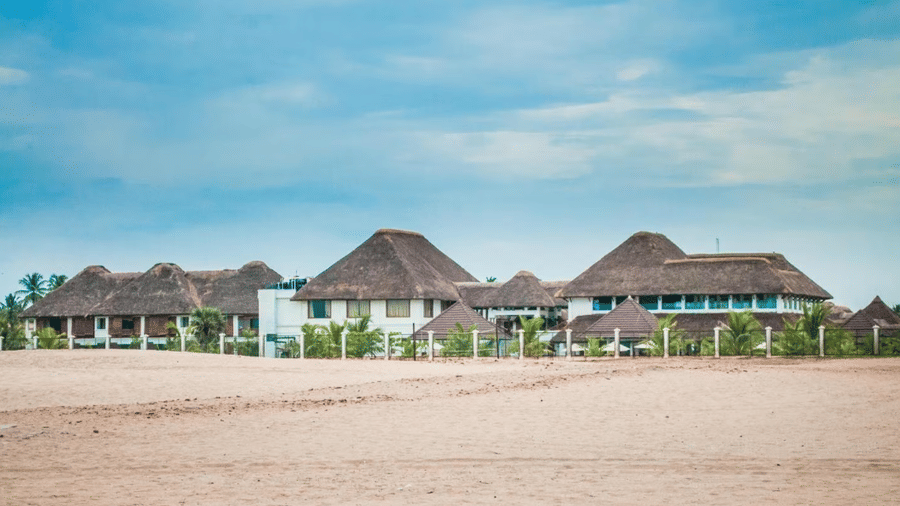 A row of resort buildings with thatched roofs situated behind a sandy beach under a blue sky | Le Pondy