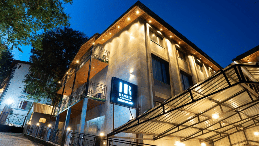 An evening view of the hotel exterior with warm lighting, glass awnings, and a blue illuminated resort sign | Lawrence Solitaire Resort, Nainital