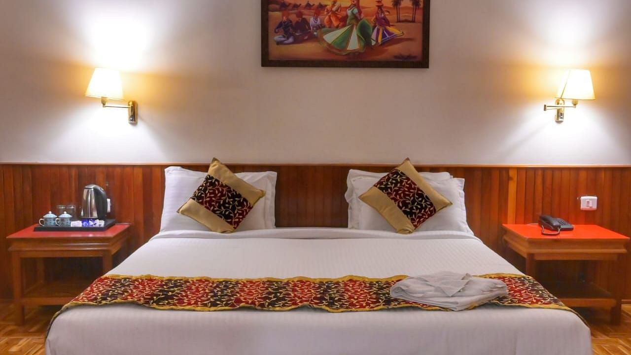 Warmly lit hotel room featuring a double bed with patterned cushions and runner, bedside tables with lamps, a tea kettle setup, and a traditional painting above the headboard - Sumitel Chiminda, Pelling.