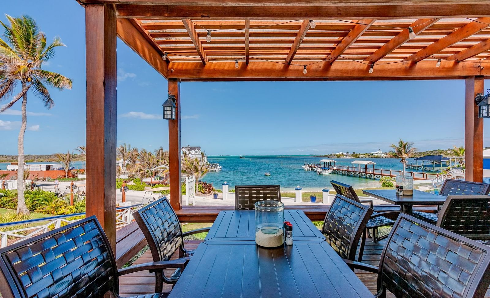 Outdoor dining setup under a wooden pergola with a view of palm trees, boats, and water in the background.