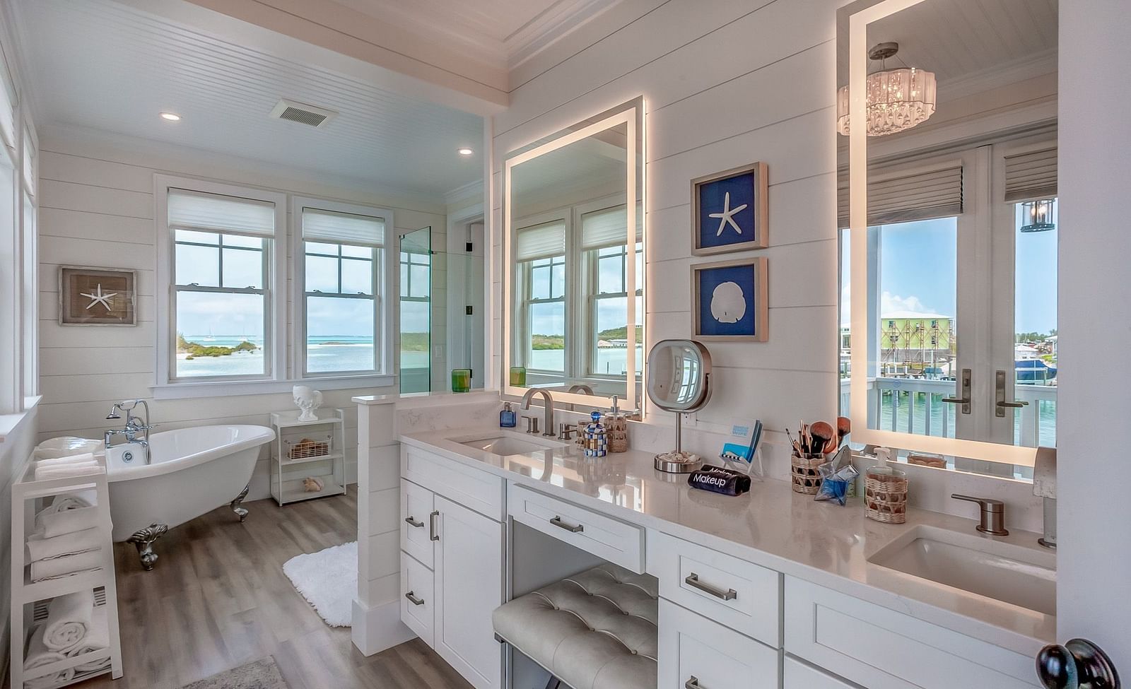 a bright and airy bathroom with a bathtub, shelves with towels, a makeup area with two huge mirrors, drawers and a chair with windows that overlooks the sea at Abaco Inn.