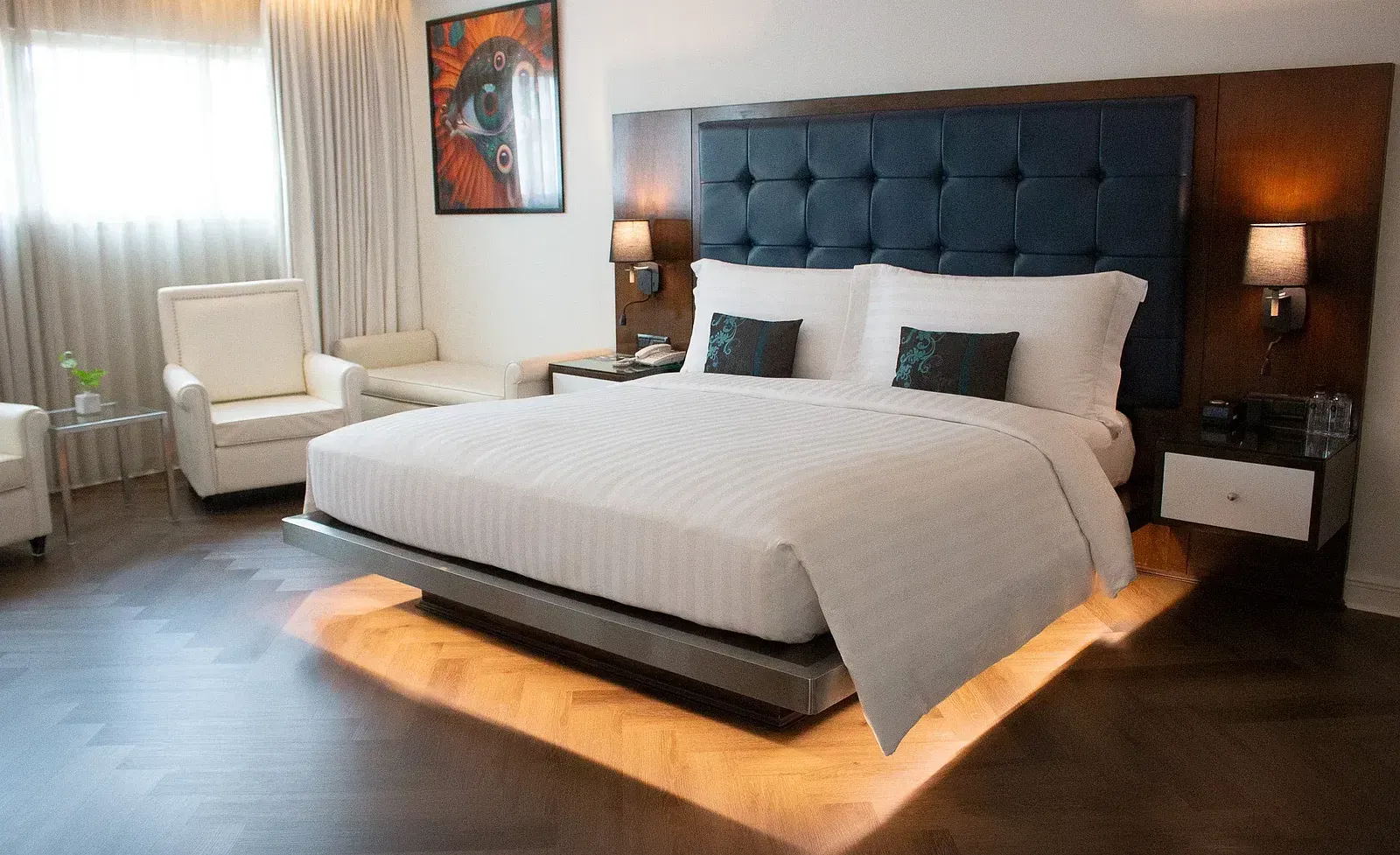 Luxurious hotel suite bedroom with illuminated platform bed, tufted headboard, vibrant artwork, cream armchairs, and warm ambient lighting throughout at Night Hotel Bangkok.
