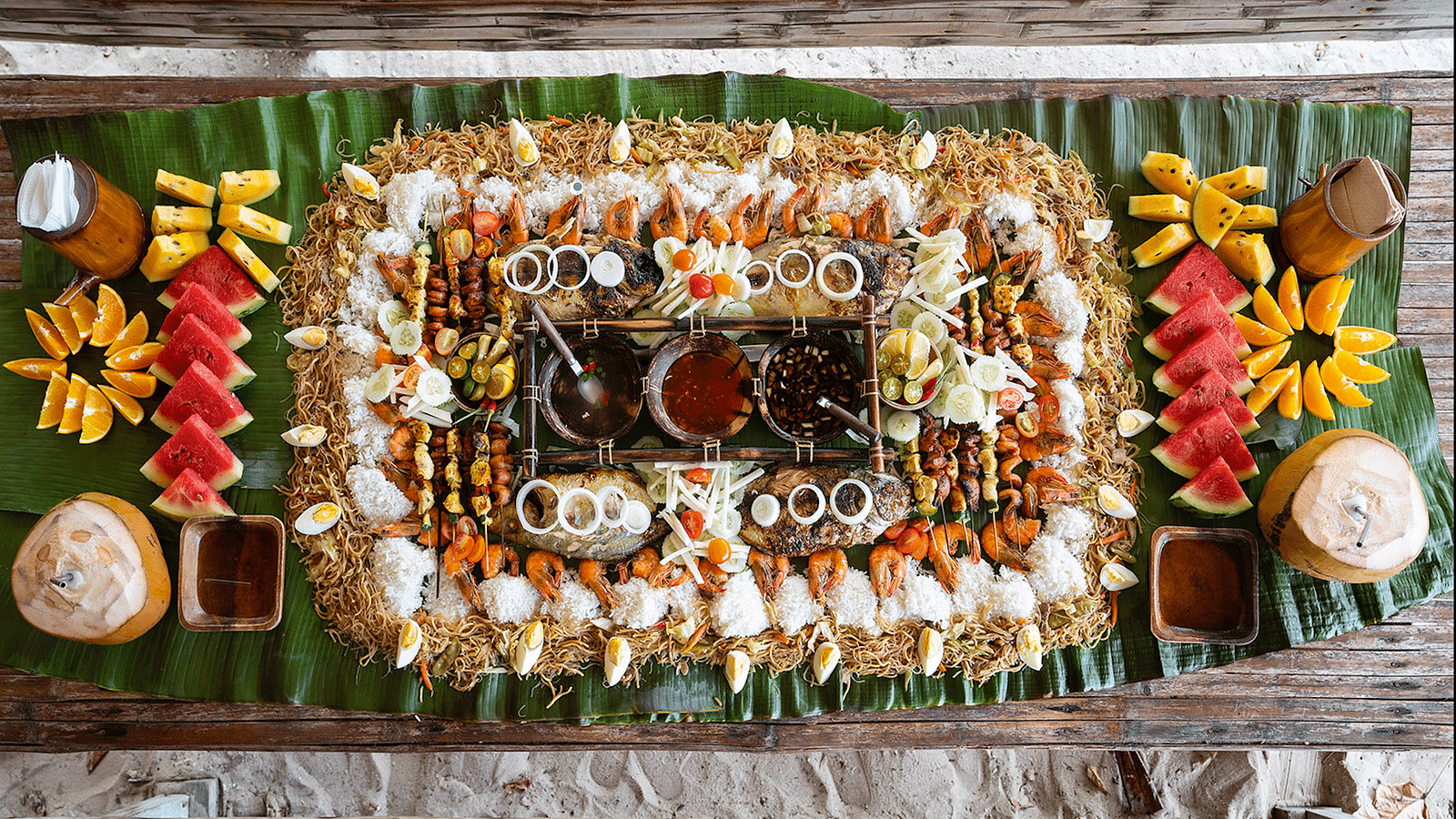 Table with assorted seafood, fruits, and rice dishes arranged on leaves at Flower Island Resort.