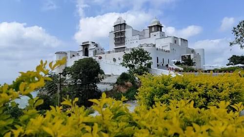Sajjangarh Monsoon Palace, rises above manicured gardens and vibrant yellow foliage, set against a partly clouded blue sky.