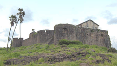 A stone fort wall with round bastions on a hill with a coconut trees beside it.