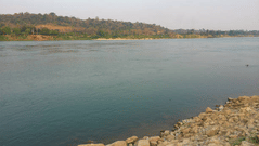 A wide river stretches to the horizon, bordered by a rocky shoreline and a distant forested hill under a pale sky.