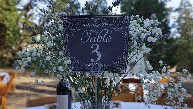 A wedding reception table is set outdoors with a 'Table 3' sign, floral arrangements, and a bottle of wine