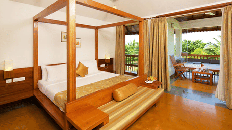 A hotel room featuring a wooden 4-poster bed and an open doorway leading out onto a private balcony with outdoor seating | Le Pondy