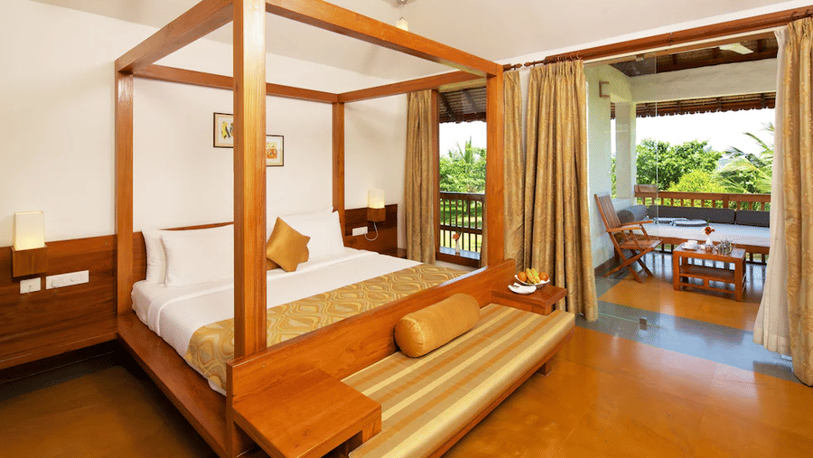 A hotel room featuring a wooden 4-poster bed and an open doorway leading out onto a private balcony with outdoor seating | Le Pondy