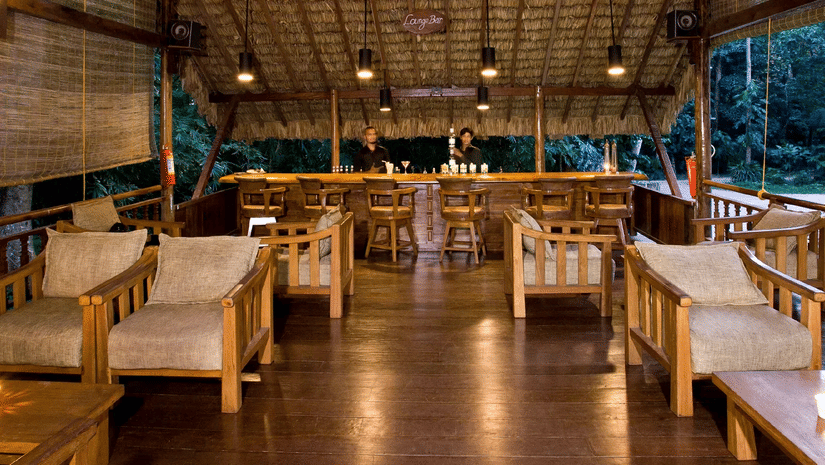 A spacious open-air wooden seating area with sofas and tables, featuring a high ceiling and large openings that overlook trees and greenery near Neil Cove Beach, Havelock, at Barefoot at Havelock.