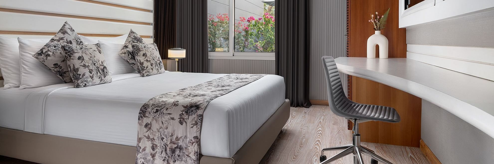 A chic room at The Atara, Gurugram with light wood flooring, a neatly made bed, a spacious desk with an office chair, and ample natural light from a large window
