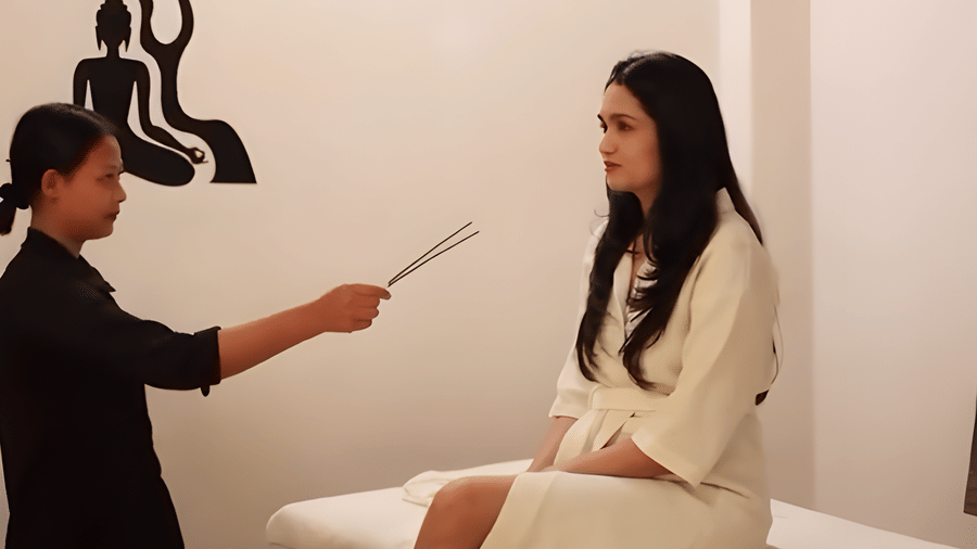 A woman sitting on the spa bed while a spa specialist stands before her holding incense sticks at Bamboo Saa Sunrise Wedding & Wellness Resort, Jaipur.