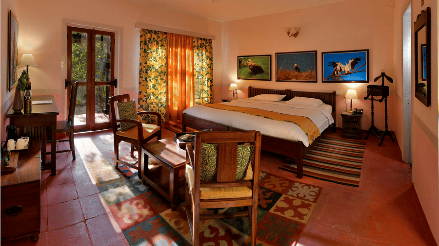 Bright heritage bedroom with wooden bed seating area patterned floor rugs artwork and long windows allowing natural light throughout the room at Neemrana’s Glasshouse on The Ganges offering a welcoming and restful environment