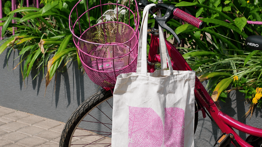 A tote bag hangs from the handlebar of a bicycle parked next to green bushes at Bougainvillea, Alibaug.