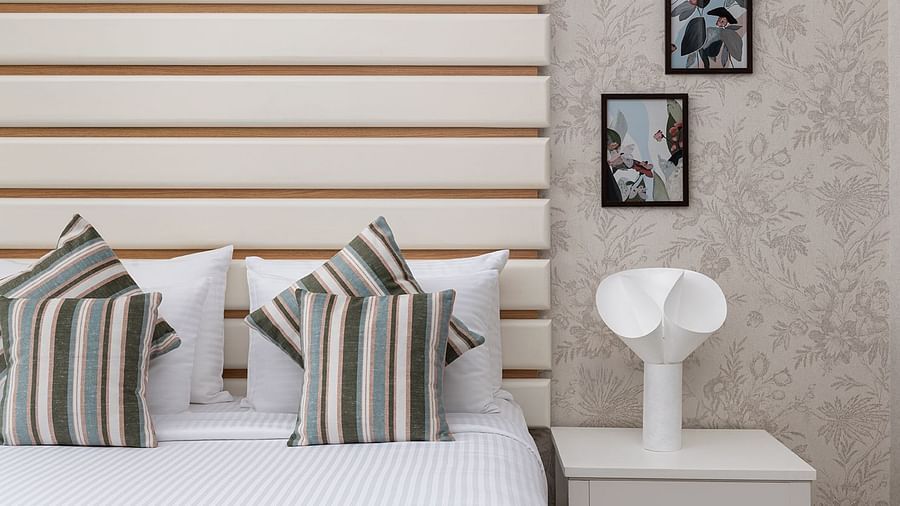 A minimalist bedroom at The Atara, Gurugram featuring a neatly made bed with white linens and striped pillows, accented by a light wood headboard with horizontal slats and framed artwork