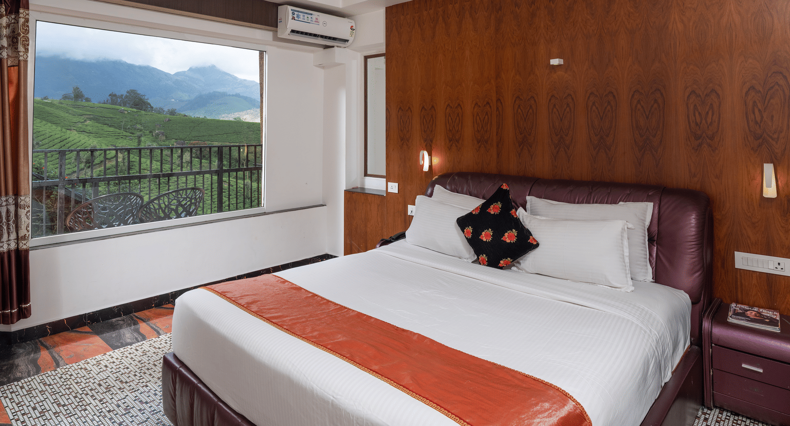 The bedroom of Premium Room at Parakkat Nature Hotel and Resorts, Munnar featuring a large bed, textured wood panelling on the wall, air conditioning, and a stunning view of the hilly landscape through a window.