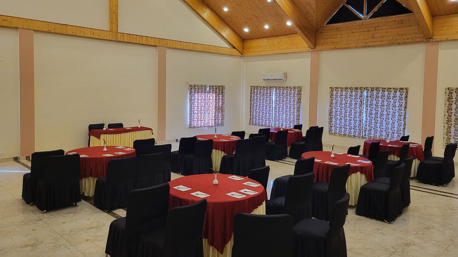 A spacious banquet hall with wooden ceiling beams, round tables, and elegant black and red seating at Dagshai Manor Kasauli - Inde Hotels.