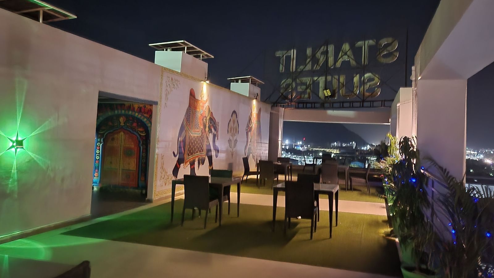 Outdoor rooftop dining at Starlit Suites, Neemrana, at night. Features green turf, tables, elephant mural, and city lights in background.