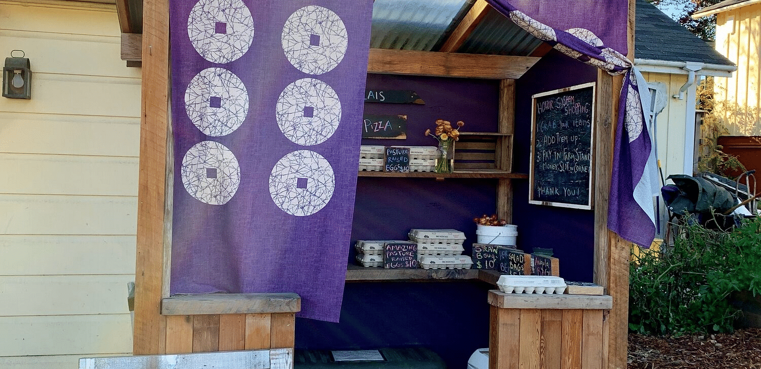 A rustic outdoor stand with a draped fabric, signage, and various items for sale on a bright day.