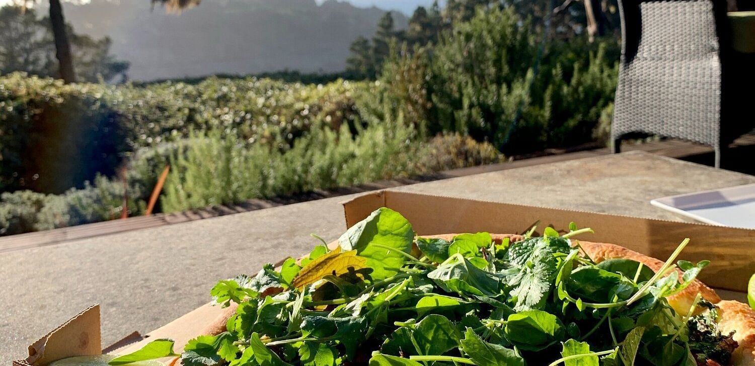 Arugula-topped pizza in an open box on a patio with a scenic view in the background.