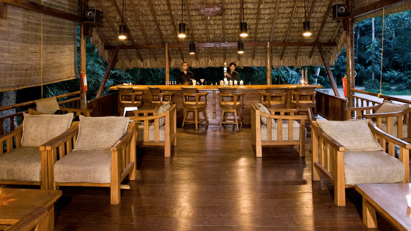 A spacious open-air wooden seating area with sofas and tables, featuring a high ceiling and large openings that overlook trees and greenery near Neil Cove Beach, Havelock, at Barefoot at Havelock.