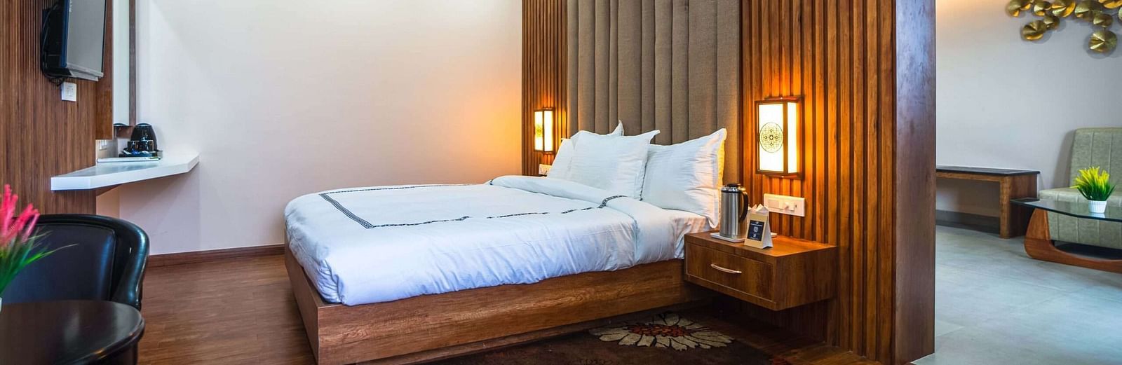 A hotel room at Mansha Palace features a bed, nightstands with lamps, a flat-screen TV, a ceiling fan, and a partial view into a seating area.