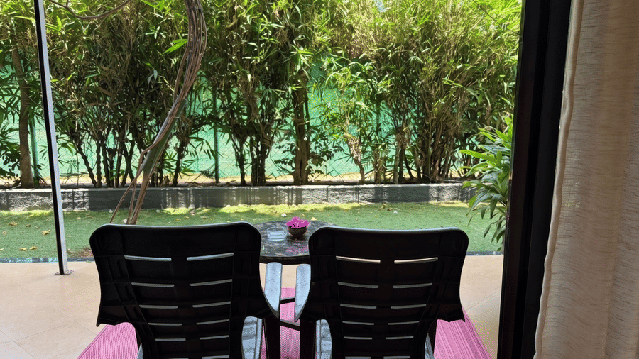2 chairs and a small coffee table sit on a pink mat on a balcony overlooking a garden with dense hedges at Bougainvillea, Alibaug.