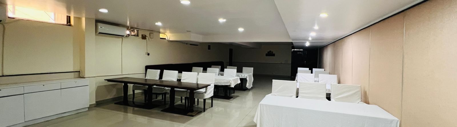  A hotel conference room with long tables and white chairs arranged for an event, featuring modern lighting and light-colored walls. | Hotel Aadhar