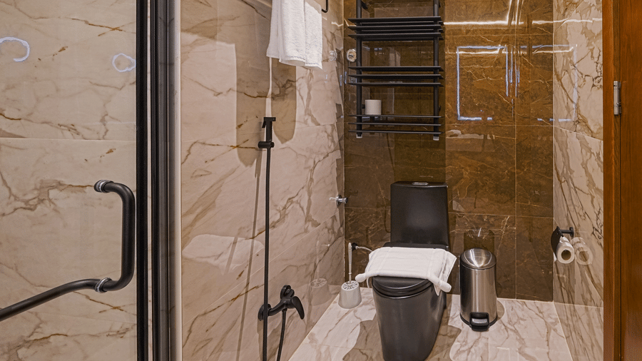 Bathroom of a Deluxe King Room at Grand Iva Hotel, Abha, featuring a black western toilet, wall-mounted shelves with towels, and tiled walls.