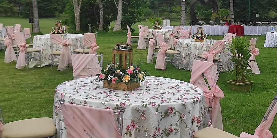 Outdoor dining setup on a lawn with round tables covered in floral tablecloths, chairs decorated with pink sashes, and a flower arrangement centrepiece - Black Thunder, Coimbatore