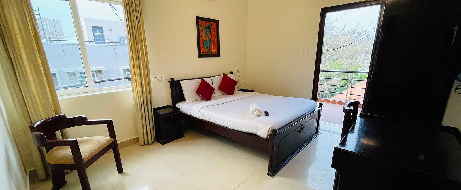 A well-lit room showcasing a double bed with white linens and red cushions, an armchair, a window with natural light, and a doorway leading to the balcony at Sanctum Suites Indiranagar, Bangalore.