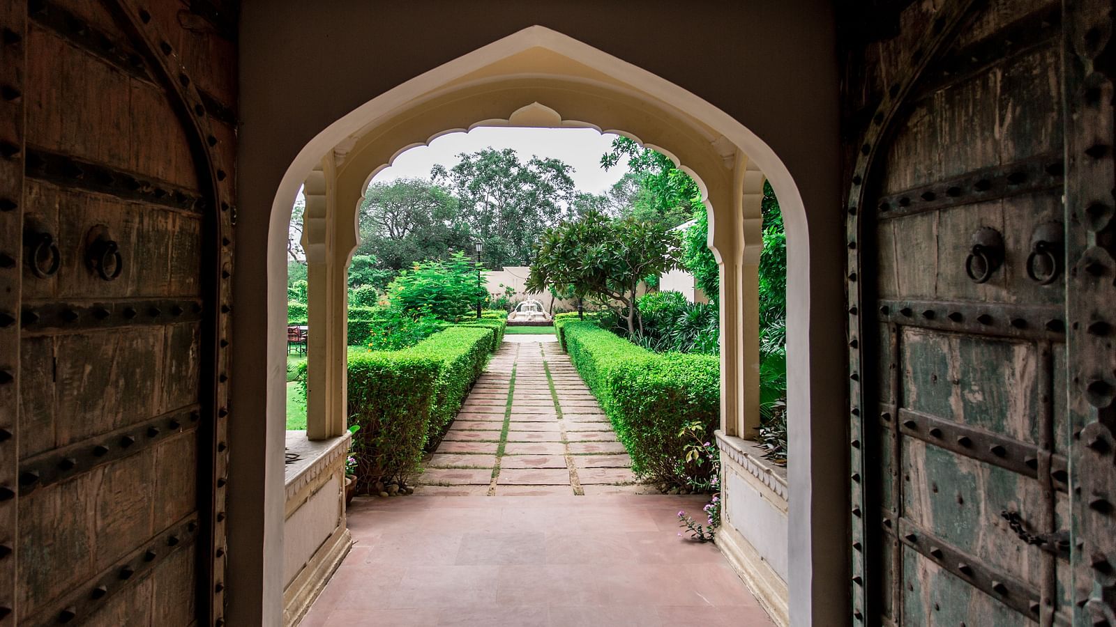 An arched doorway offering a view of a long, hedged pathway leading away from Royal Heritage Haveli.