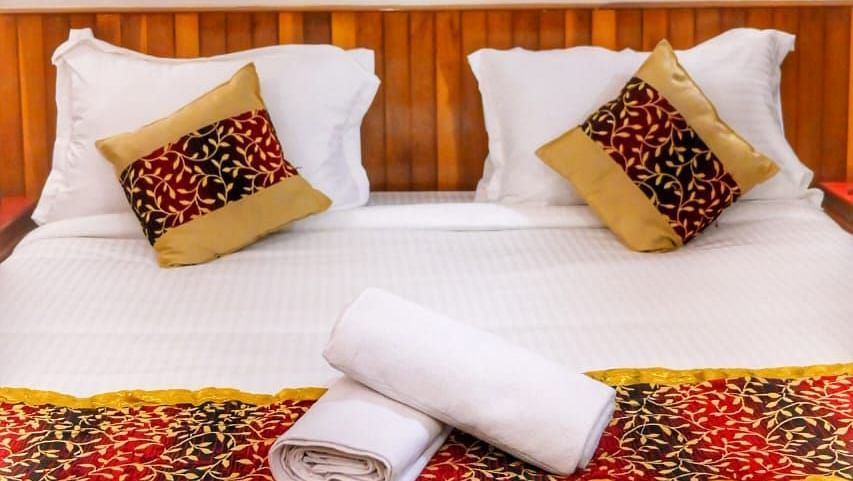 Neatly made double bed with traditional decorative cushions and runner, white linens, and rolled towels, set against a wooden headboard and wall art depicting a cultural Indian painting - Sumitel Chiminda, Pelling.