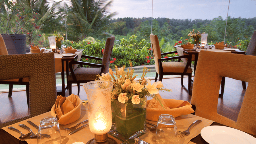 Candle-lit dining tables with plates, cutlery, napkins and floral centrepieces placed indoors, facing large glass windows that overlook greenery and palm trees at The Serai Chikmagalur.
