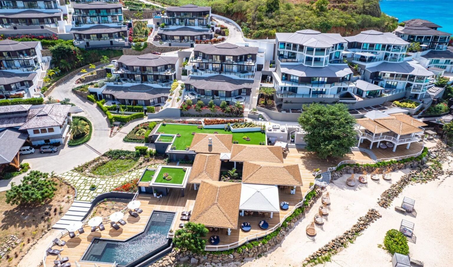 An aerial view of a luxury resort complex with multiple buildings, pools, and lush landscaping, situated near a coastline.