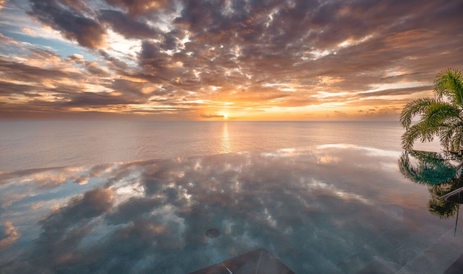 A serene tropical sunset over the ocean with a reflection of the sky on the water and a palm tree visible on the right.