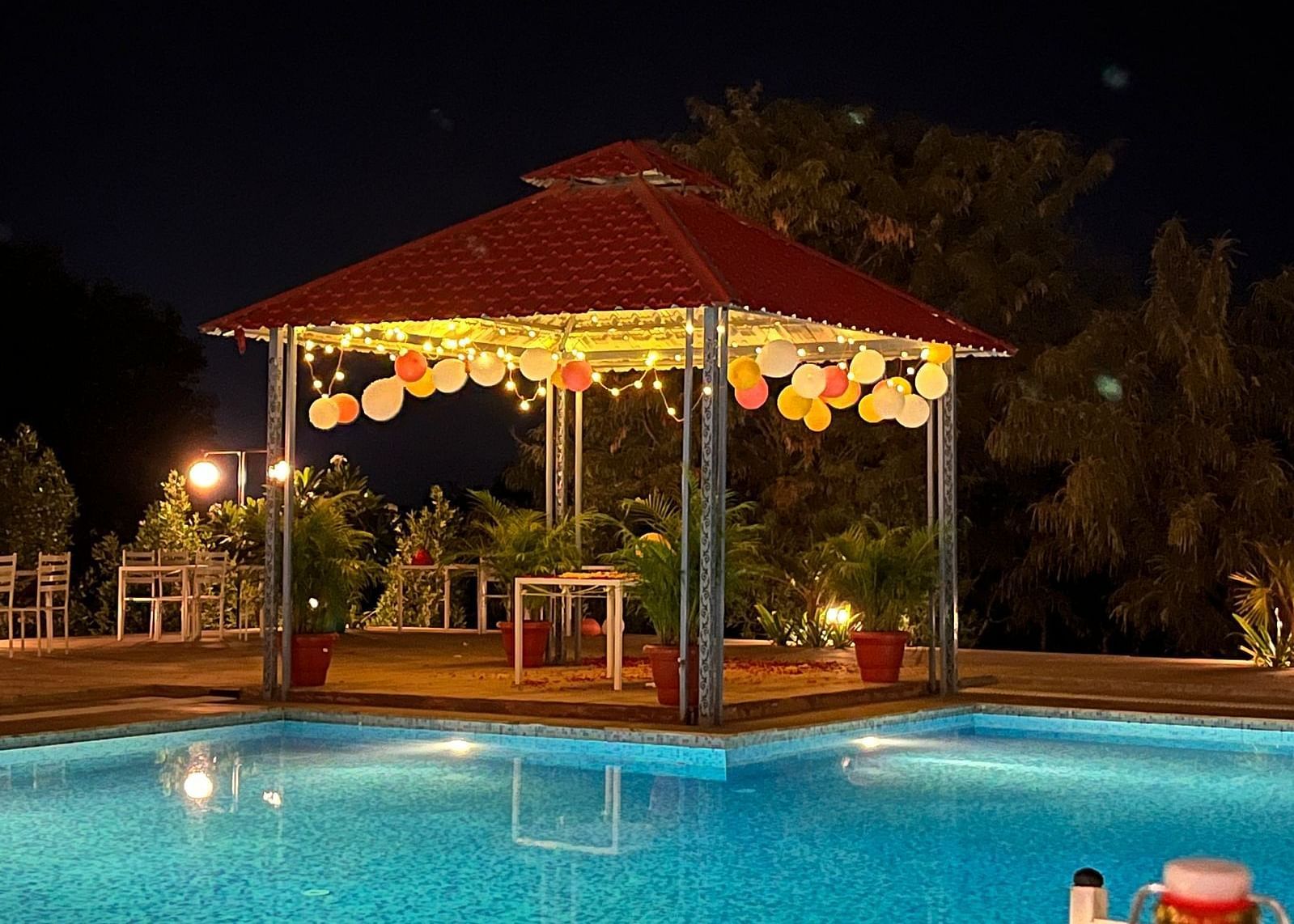 A nighttime shot of a gazebo with a red roof, decorated with lights and balloons, next to a brightly lit swimming pool - Bamboo Saa Mulberry Resort, Pushkar